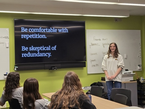 A young woman with wavy, light brown hair stands next to a TV in the Law Writing Center. She is presenting to a group of students who are seated at a table. On the TV, there's white text against a black background. The text says, "Be comfortable with repetition. Be skeptical of redundancy."